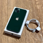 Green iPhone with dual camera back, resting on its white box on a wooden floor, with a coiled white charging cable nearby.