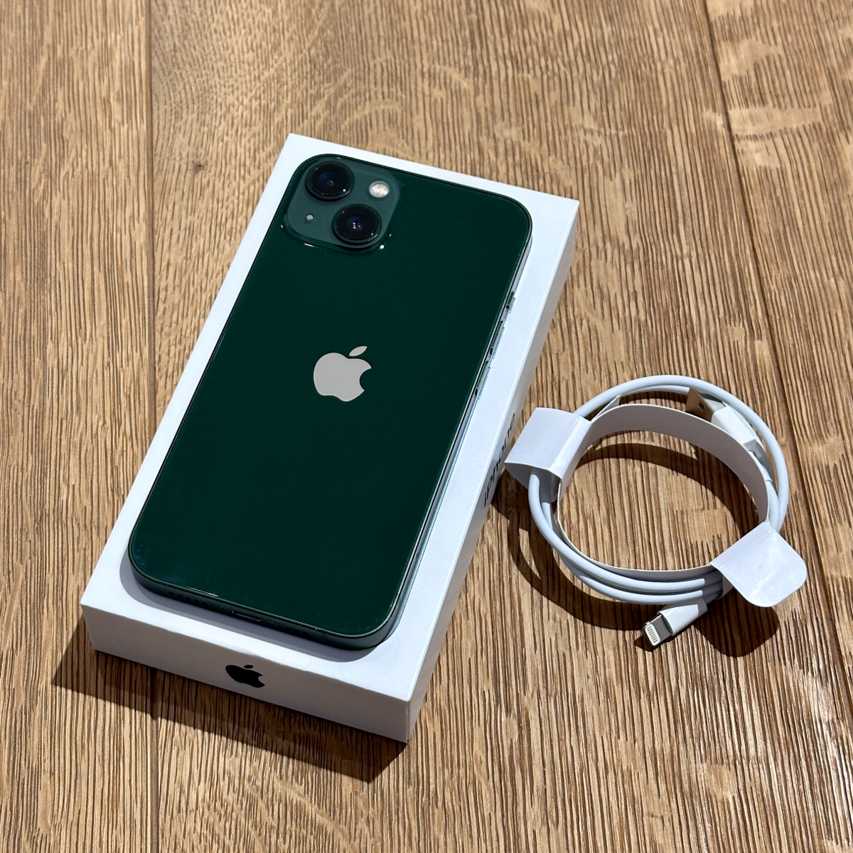 IMG_9619 Green iPhone with dual camera back, resting on its white box on a wooden floor, with a coiled white charging cable nearby.