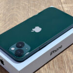 Green iPhone with a dual-camera system on the back, resting on its white box showing the iPhone label.