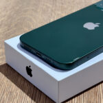 Green iPhone resting on its white box, angled to show the bottom edge with speaker holes and Lightning port.