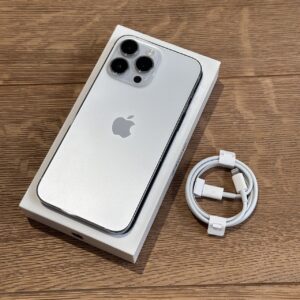 Silver iPhone with triple-camera back in its box on a wooden surface, next to a coiled white charging cable.