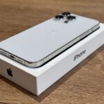 White iPhone resting on its box, showing a triple-camera back and Apple logo on a wooden surface