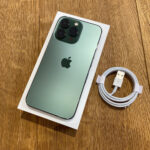 Mint green iPhone with triple-camera back on its white box, placed on a wooden floor with a white USB-A to Lightning cable beside it.