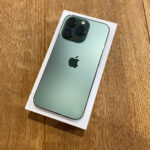 Mint green iPhone viewed from the back, showing triple-camera setup on the top left, placed on its white box on a wooden surface.