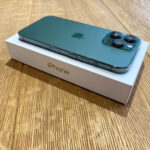 Teal iPhone 13 Pro on its white box, showing the triple-camera setup on the back, angled on a wooden table.