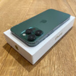 Green iPhone on its white box, showing a triple-camera system and Apple logo on a wooden surface