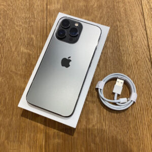 Silver iPhone with triple-camera back in its box on a wooden table, USB charging cable beside it.