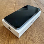 Black iPhone resting face up on its white Apple box on a wooden table, with the device’s screen off.