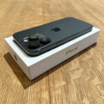 Black iPhone resting on its white box, rear triple-camera setup visible on a wooden surface