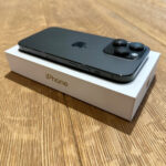 Black iPhone 15 Pro on its white box, viewed from a top-right angle on a wooden surface, showing triple-camera module and Apple logo.