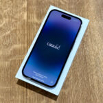 Smartphone in a white box resting on a wooden surface; the screen shows a blue-to-purple gradient wallpaper.