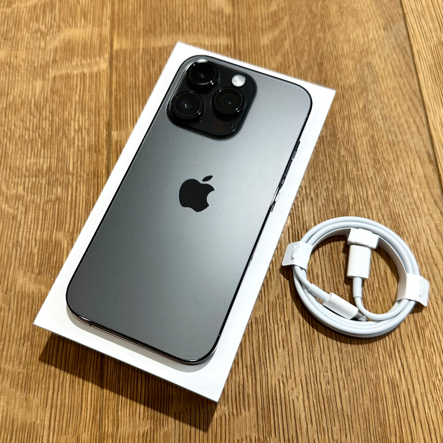 Grey iPhone with triple-camera setup resting on its white box on a wood surface, with a coiled white charging cable nearby