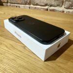 Black iPhone with triple-camera back resting on a white box on a wooden table, brick wall in the background (product photo).