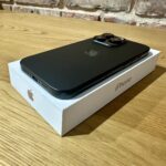 Black smartphone with a triple-camera rear setup resting on its white box on a wooden table, brick wall in the background as scenery.