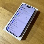 iPhone visible in its white box on a wooden table, screen showing the Settings app with menu items like Apple Account and Wi‑Fi