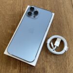 Light blue iPhone with triple-camera setup resting on a white box, next to a coiled white charging cable on a wooden surface.