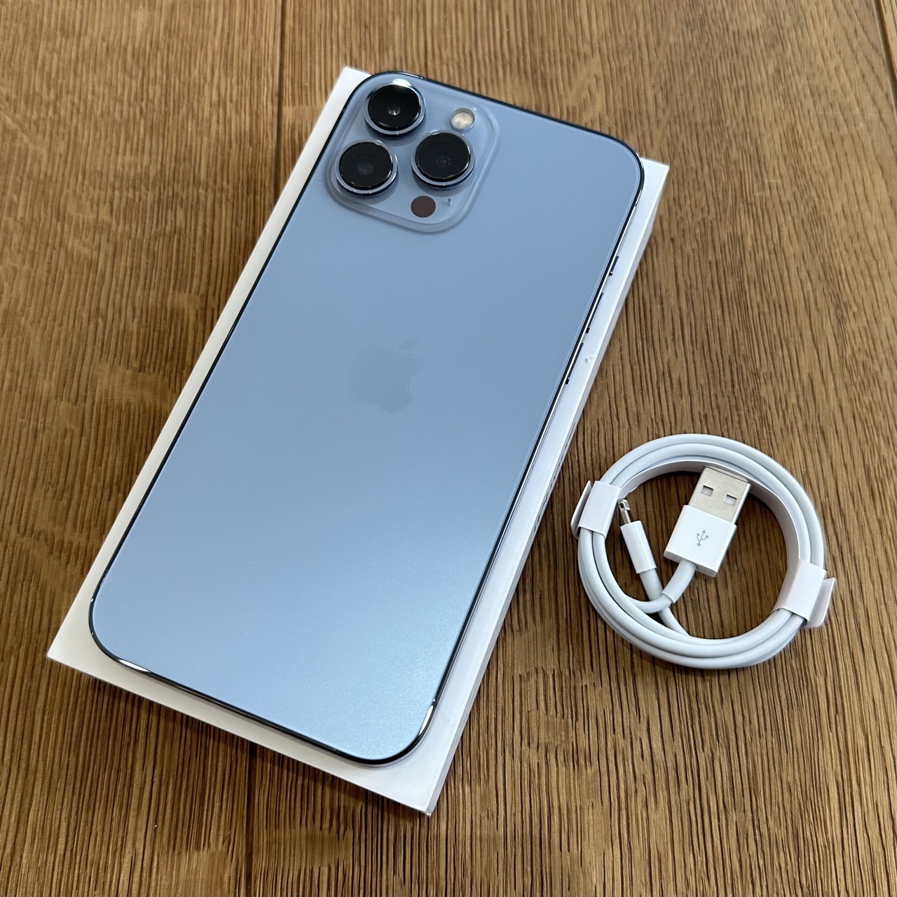 Light blue iPhone with triple-camera setup resting on a white box, next to a coiled white charging cable on a wooden surface.