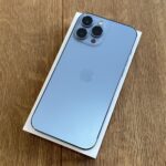 Blue iPhone with a triple-lens camera back, resting on its white box on a wooden surface.