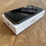 Black iPhone resting on its white box labeled iPhone, on a wooden table with a glossy screen off.