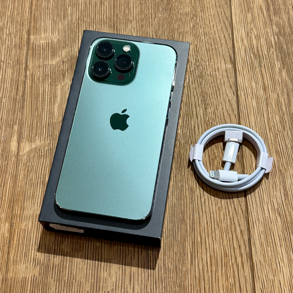 Turquoise-green iPhone in its box on a wooden floor, with a coiled white USB-C to Lightning cable beside it