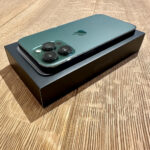 Teal iPhone with three-camera back resting on its box on a wood table.