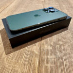 New green iPhone with triple-camera setup resting on its black iPhone box on a wooden table, angled to show the back and cameras.