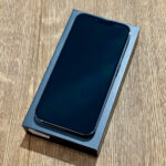 Black smartphone with an edge-to-edge screen resting on its blue retail box on a wooden table.