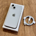 Silver iPhone in its box on a wooden surface with a coiled white charging cable beside it.
