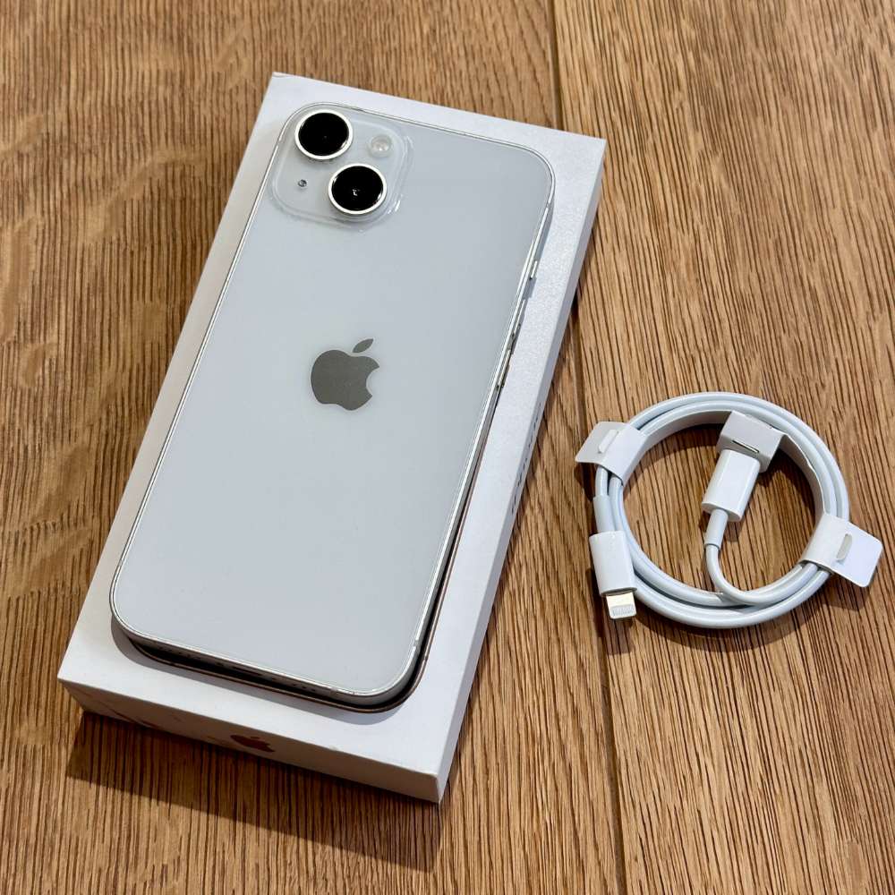 Silver iPhone in its box on a wooden surface with a coiled white charging cable beside it.