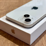 White iPhone with dual-camera module placed on its box on a wooden surface, Apple logo visible in the background.