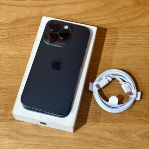 Black iPhone with triple-camera array on its box, resting on a wooden table beside a braided white charging cable.