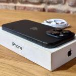 Black iPhone with triple-lens camera resting on its white box on a wooden table behind.