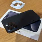 Black iPhone with triple-camera setup lying on its box on a wooden surface; white USB-C charging cable in the background
