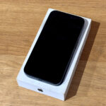 Black iPhone in its white Apple box resting on a wooden table, screen off on display.