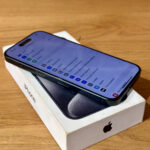 Black iPhone resting on its white box on a wooden surface, screen angled upward showing app icons on a blue tint.