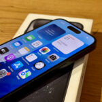 iPhone with a blue iOS home screen showing calendar and app icons on a wooden table surface.