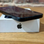 Black smartphone resting on a white Apple box on a wooden table, showcasing the bottom edge and logo visible on the box.