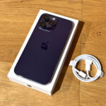 Purple iPhone with triple-camera back, in its white box on a wooden surface, with a coiled white charging cable nearby.