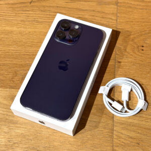 Purple iPhone with triple-camera back, in its white box on a wooden surface, with a coiled white charging cable nearby.
