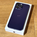 Purple iPhone with triple-camera array in its white box, lying on a wooden table, viewed from above.
