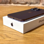 Purple iPhone resting on its white box on a wooden table, showing the bottom edge with ports and speaker grills.