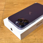 Purple iPhone with triple-camera back resting on its white box on a wooden surface, showing the rear camera array and Apple logo.