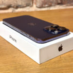 Purple iPhone with triple-camera system resting on its white packaging box on a wooden table surface.