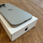 Silver iPhone with a triple-camera back resting on a white box on a wooden floor, Apple logo visible on the back.