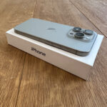 Boxed silver iPhone on a wooden table, showing the Apple logo and three camera lenses in the upper corner.