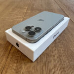 Silver iPhone box on a white box, showing the phone’s triple-camera module on a wooden table.